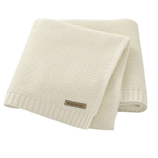 A folded knitted baby blanket in a light color, displayed on a textured surface.