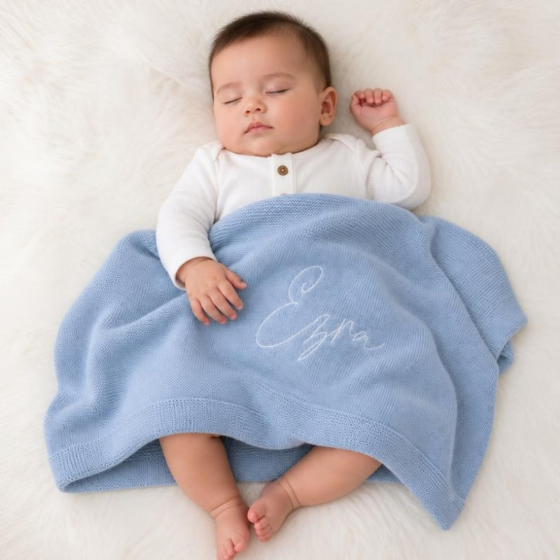 A sleeping baby wearing a white long-sleeve top, wrapped in a light blue knitted blanket with the name 'Ezra' embroidered in white script, lying on a white faux fur surface.