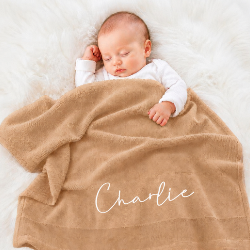 A sleeping newborn baby wrapped in a soft tan blanket with the name 'Charlie' embroidered in white script, lying on a white faux fur rug.