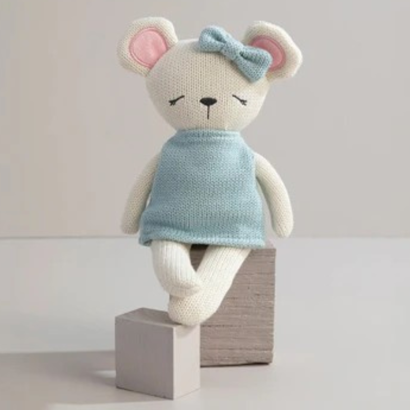 A knitted mouse doll with cream-colored fur, pink inner ears, a light blue knitted top, and a matching bow on its head, sitting on two small blocks against a plain light background.
