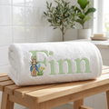 White towel with embroidered name in pale green and peter rabbit character on a wooden bench in a bright room.