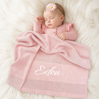A sleeping baby wrapped in a soft pink blanket with the name 'Eden' embroidered in white script, lying on a white faux fur surface. The baby wears a pink long-sleeve top and a pink daisy headband.