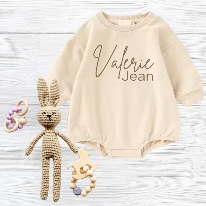 Beige baby romper embroidered with 'Valerie Jean' text, knitted bunny, and wooden toys on white wooden background