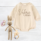 Beige baby romper embroidered with 'Valerie Jean' text, knitted bunny, and wooden toys on white wooden background