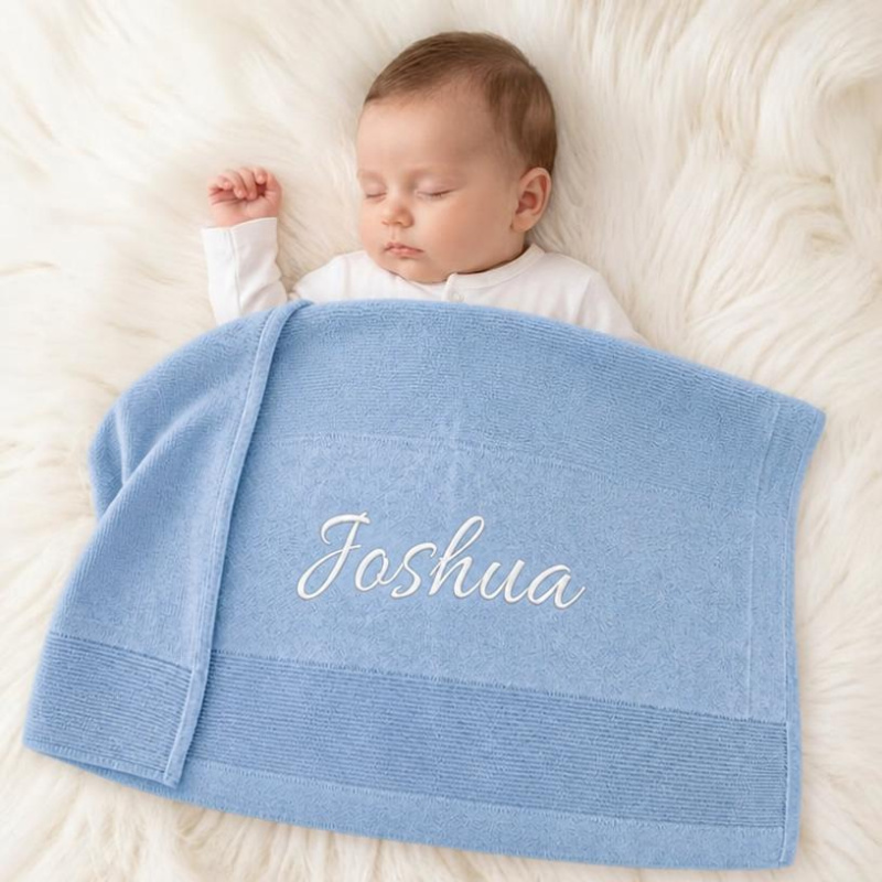 A sleeping baby wrapped in a light blue personalized blanket with the name 'Joshua' embroidered in white script, lying on a white faux fur surface.