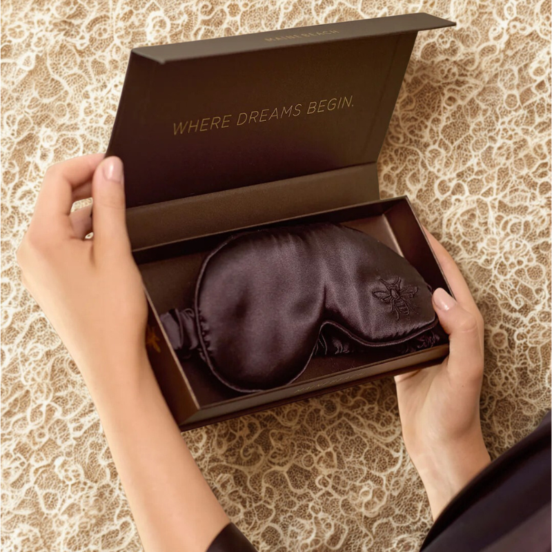 Brown satin eye mask in an open box with 'Where Dreams Begin' text on a patterned background.