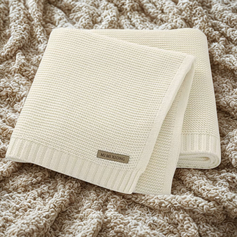 A folded knitted baby blanket in a light color, displayed on a textured surface.