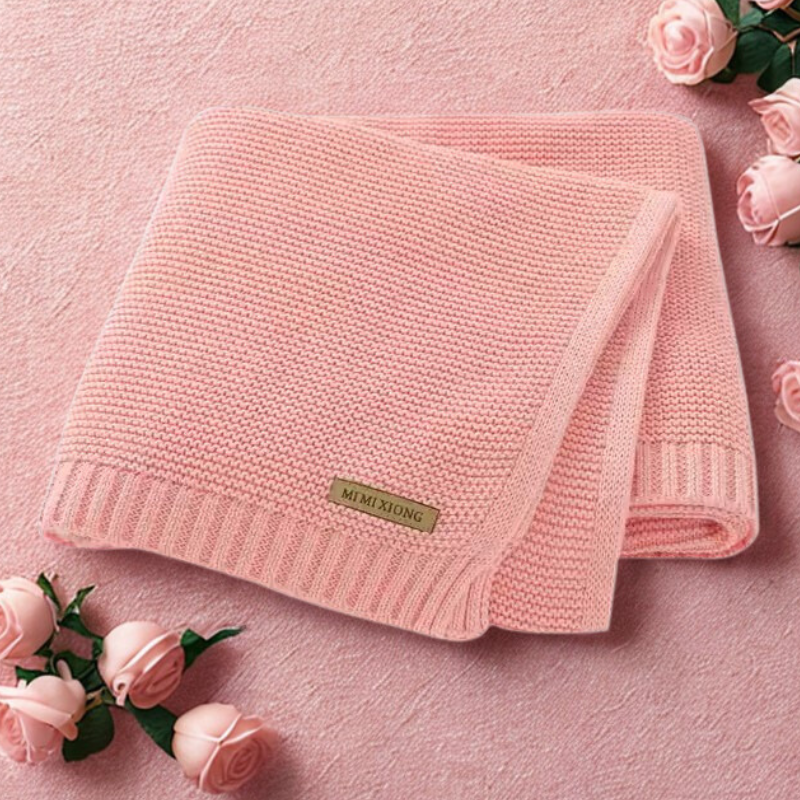 Pink knitted blanket with a label on a pink surface surrounded by pink roses.