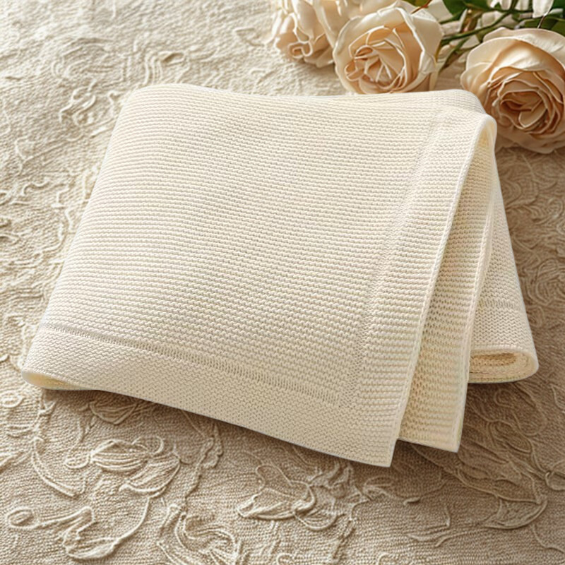 A folded beige knitted baby blanket with a solid line bordered edge, displayed on a similar beige fabric with floral patterns, with two roses at the top of the image.