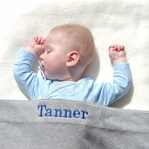 Baby in a blue outfit lying on a white blanket with a gray knitted blanket personalised with the name 'Tanner'.