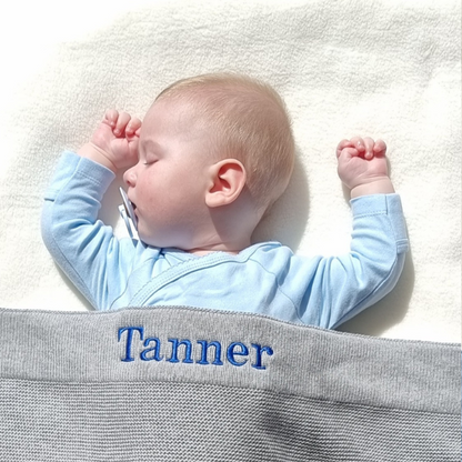 Baby in a blue outfit lying on a white blanket with a gray knitted blanket personalised with the name 'Tanner'.