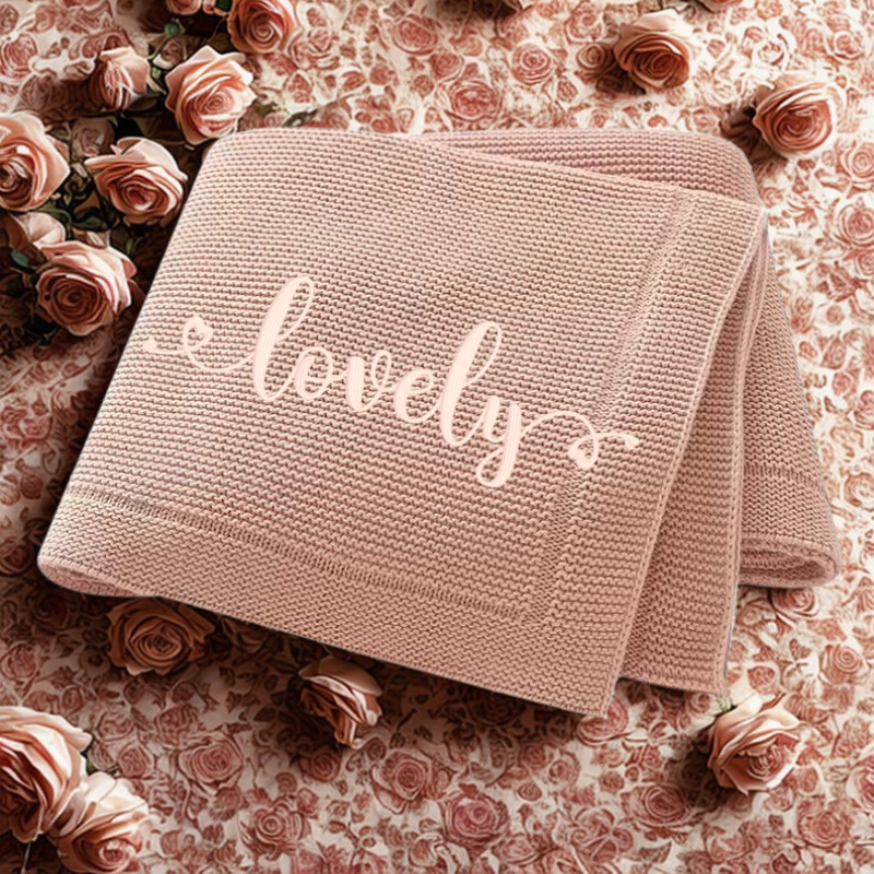 Pink knitted baby blanket with 'lovely' text on a floral background.