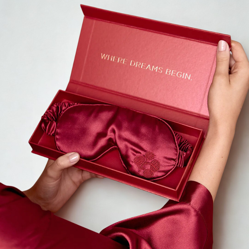 Red satin eye mask in a box with 'Where Dreams Begin' text, held by a person.