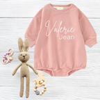 Pink baby romper with 'Valerie Jean' text, knitted bunny, and wooden toys on white wooden surface.