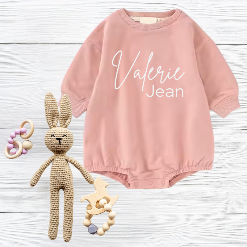 Pink baby romper with 'Valerie Jean' text, knitted bunny, and wooden toys on white wooden surface.