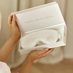 White sleep mask in a box with 'Where Dreams Begin' text, held by a person.