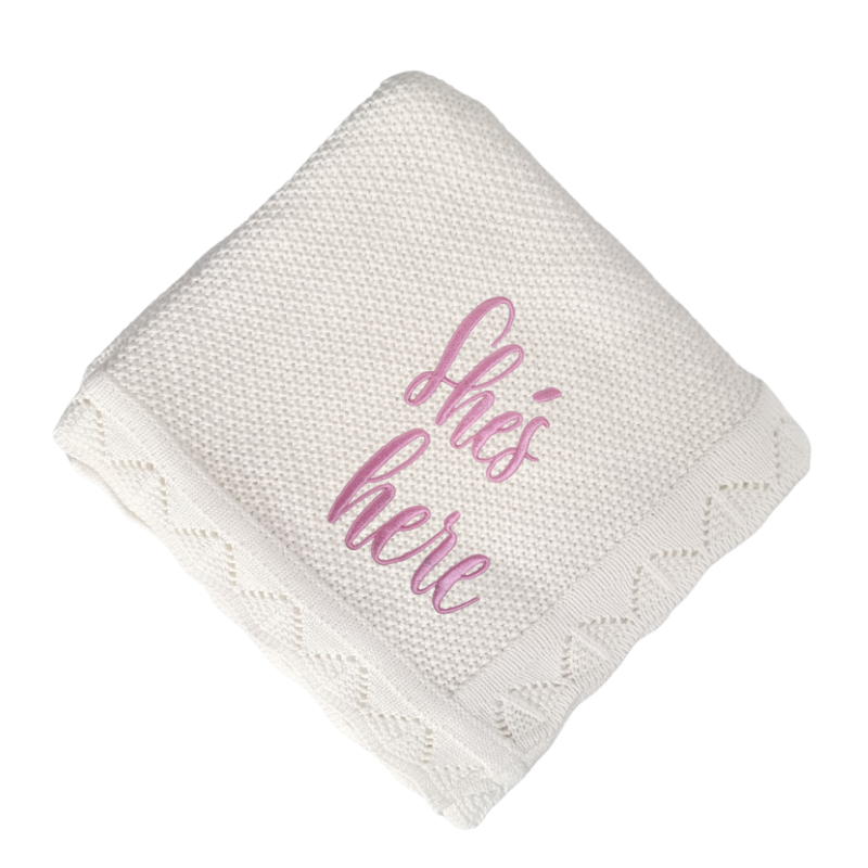 White textured blanket with pink text 'she's here' on a white background